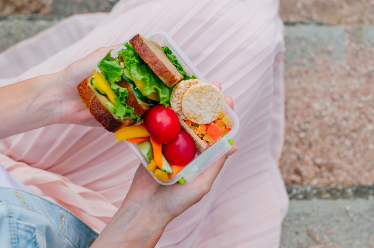 Healthy food concept: Young hipster girl holding lunch box filled with sandwich, crispbreads, fruits and vegetables sitting on stairs outdoor