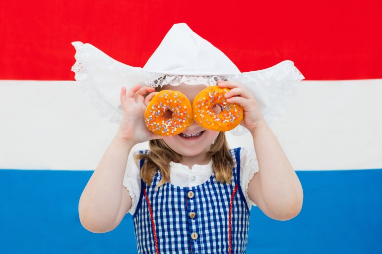 Little Dutch girl wearing traditional national costume, dress and hat holding orange donuts at flag of the Netherlands. Child with Oranje souvenirs from Holland celebrating King day. Kid with sweets.