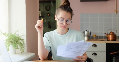 Indoor picture of young European female sitting at home at table reading data in sheets of paper with pencil in order to figure out something important and check it, feeling relaxed and confident