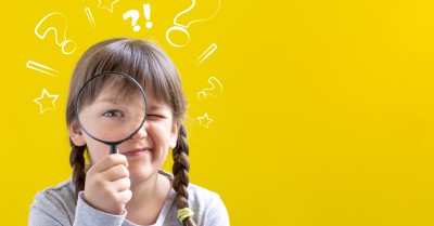 Cute curious little girl looking through a magnifying glass on yellow background with copy space.